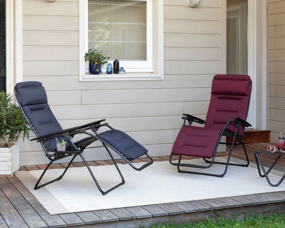 Two padded outdoor zero gravity chairs on a white patio rug in front of a beige house wall