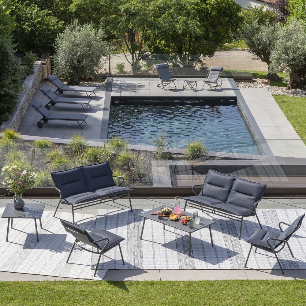 Modern outdoor patio furniture set arranged around a rectangular swimming pool in a landscaped garden. The set includes two dark gray upholstered sofas, two matching armchairs and two small side tables on a light stone patio.